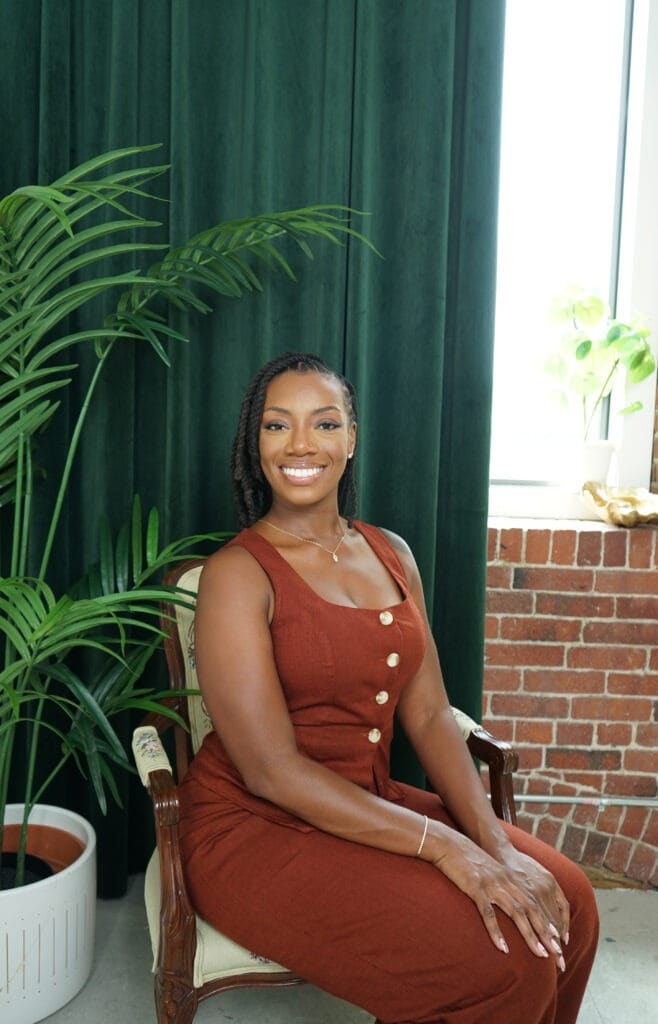 Beautiful Black woman sitting on a chair with a brown/red dress and a bright genuine smile. there is a black and curtain in the background.
