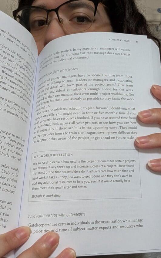 A woman with dark, curly hair and glasses smiles warmly as she holds up a copy of Elizabeth Harrin's "Managing Multiple Projects," 2nd edition. The book is open to a page showing Real World Reflection.