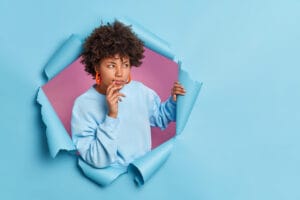 A person with curly hair wearing a light blue sweatshirt and orange earrings looks thoughtful while peeking through a torn blue paper background, revealing a pink backdrop behind them.