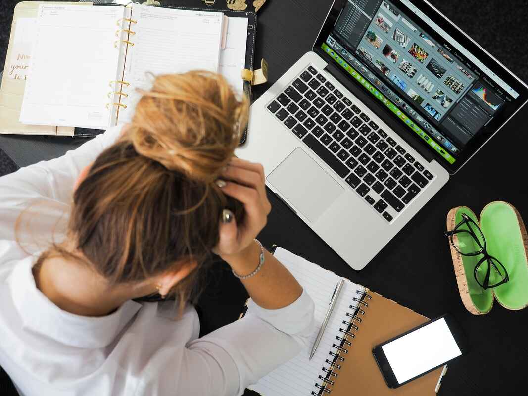 A top-down view of a person sitting at a black desk, holding their head in their hands in a gesture of stress or frustration. The desk is cluttered with an open laptop displaying a photo gallery, a large planner, a spiral notebook, a smartphone, and a pair of glasses.