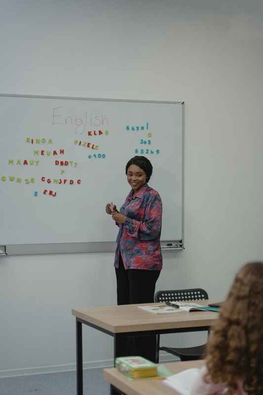 A smiling female teacher stands at the front of a classroom next to a whiteboard. The word "English" is written at the top of the board, which is also covered in colorful magnetic letters and numbers arranged in various patterns.